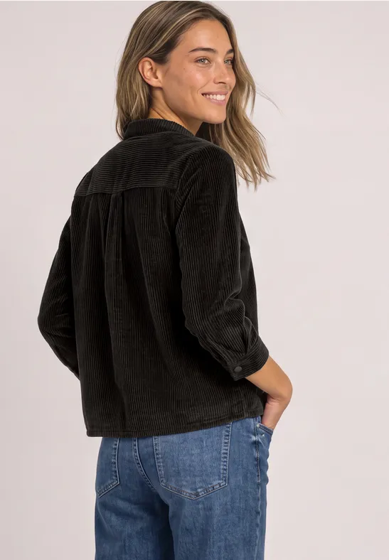 Junge Frau in schwarzer Cordjacke und blauer Jeans, lächelt und blickt über die Schulter.