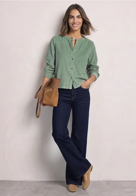 Eine Frau trägt ein grünes Cordhemd, dunkle Schlagjeans, beige Stiefeletten und hält eine Tasche vor hellem Hintergrund.