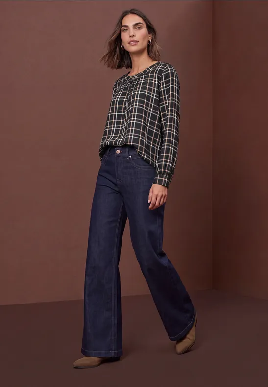 Frau in kariertem Oberteil und weiter High-Waist-Jeans vor braunem Hintergrund.