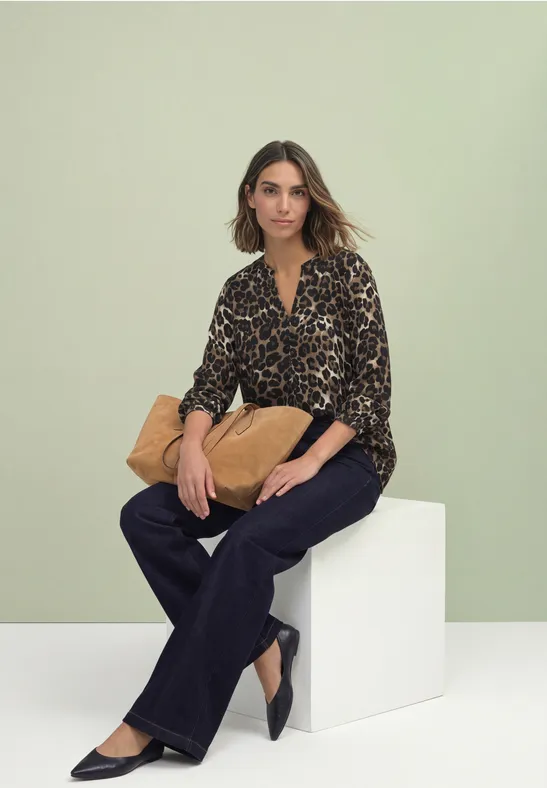 Eine Frau sitzt vor grünem Hintergrund auf weißem Block, trägt Leo-Bluse, dunkle Hose und hält eine beige Tasche.