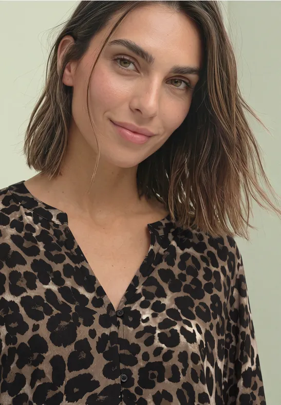 Eine lächelnde Frau mit schulterlangem braunem Haar und einem Leopardenprint-Top vor hellgrauem Hintergrund.