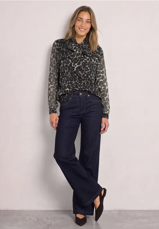 Frau trägt dunkle Schlagjeans und Animal-Print-Bluse, steht lächelnd vor hellem Hintergrund.