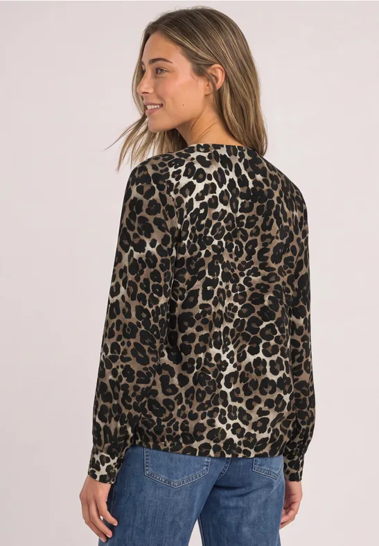Junge Frau in seitlicher Pose trägt langärmlige Bluse im Leopardenmuster und blaue Jeans vor neutralem Hintergrund.