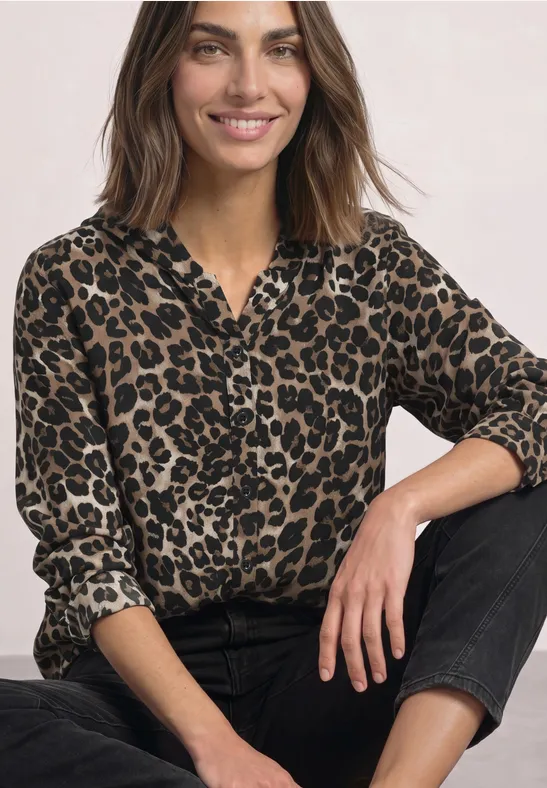 Eine lächelnde Frau mit braunen Haaren trägt eine Leoparden-Bluse und schwarze Hose vor hellem Hintergrund.