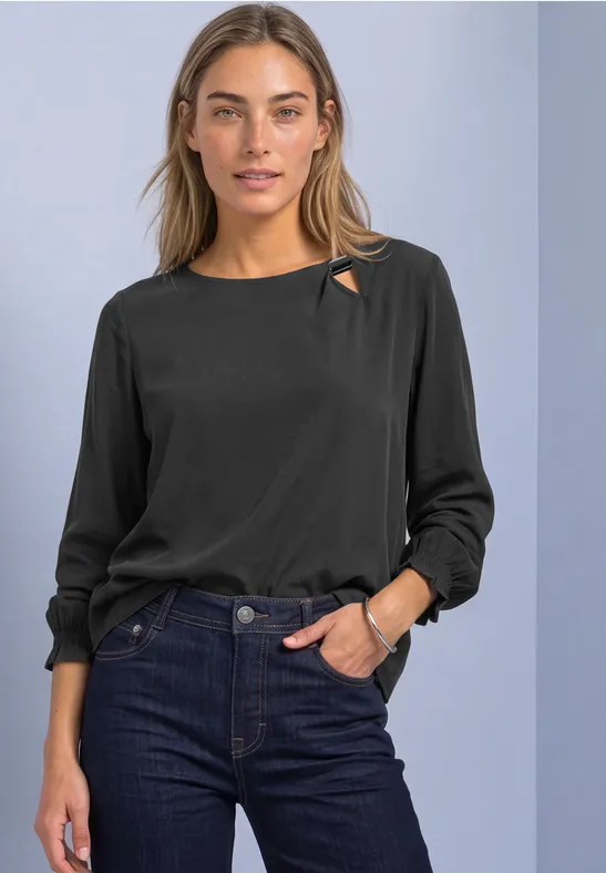 Frau in schwarzer Bluse mit Cut-out am Kragen, dunkelblaue Jeans, casual Pose vor neutralem Hintergrund.