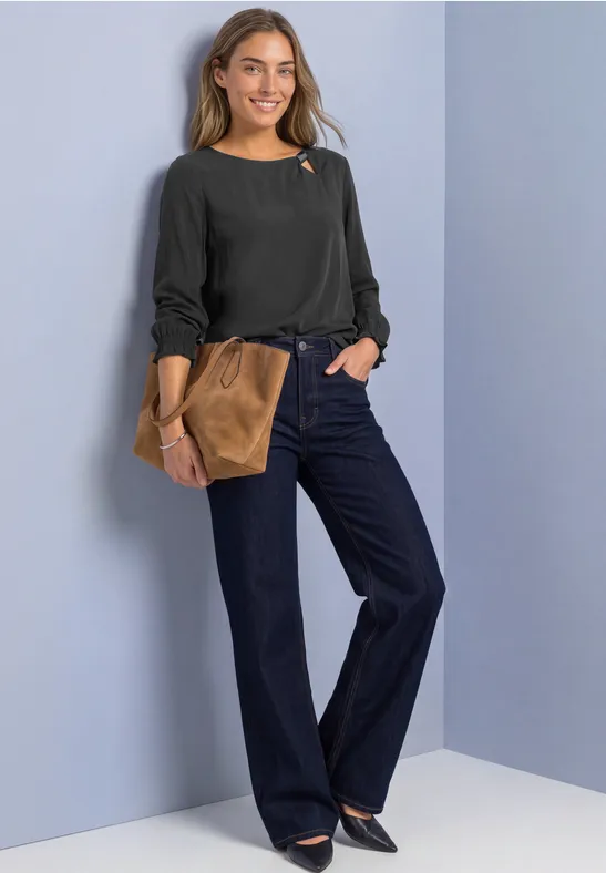 Eine lächelnde Frau in schwarzem Pullover, weiten Jeans und brauner Clutch vor hellem Hintergrund.