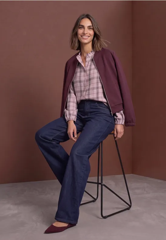 Eine lächelnde Frau sitzt lässig auf einem modernen Hocker, trägt Karobluse, weite Jeans und bordeauxfarbene Jacke.