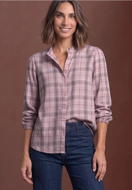 Frau mit rosa kariertem Hemd und Jeans lächelt vor braunem Hintergrund.