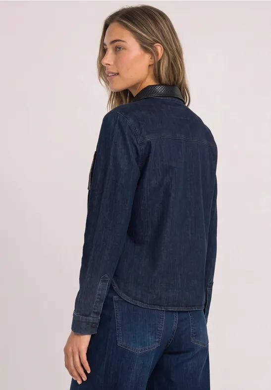 Junge Frau in dunkler Jeansjacke und Jeanshose, vor hellem Hintergrund, seitlich zur Kamera gewandt.