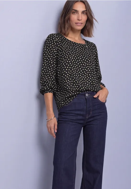 Frau in schwarzer Bluse mit weißen Punkten und weiten, dunkelblauen Jeans vor hellem Hintergrund.