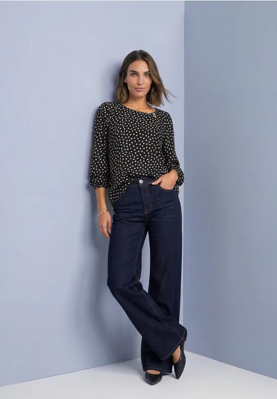 Frau in schwarzer Polka-Dot-Bluse und weiten Jeans, posiert lässig vor neutralem Hintergrund.