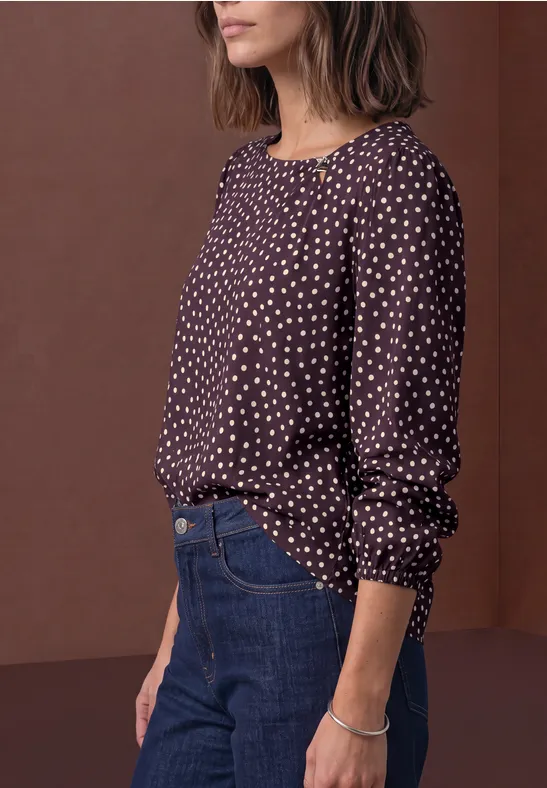 Eine Frau trägt eine dunkel gepunktete Bluse, blaue Jeans und ein Armband vor braunem Hintergrund.