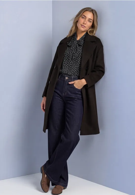 Frau in langem dunkelbraunen Mantel, gepunkteter Bluse, weiten Jeans und Stiefeletten vor schlichtem Hintergrund.