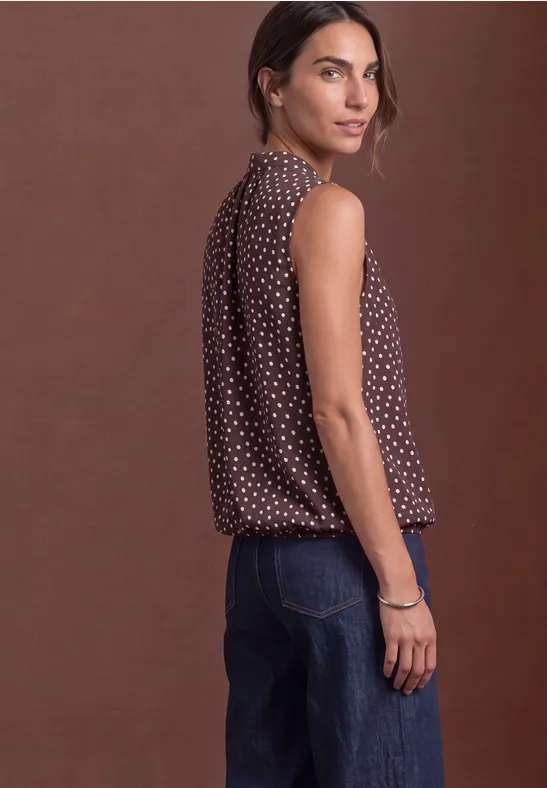 Vue de profil d'une femme portant un top marron sans manches à pois blancs et un jean devant un fond marron.