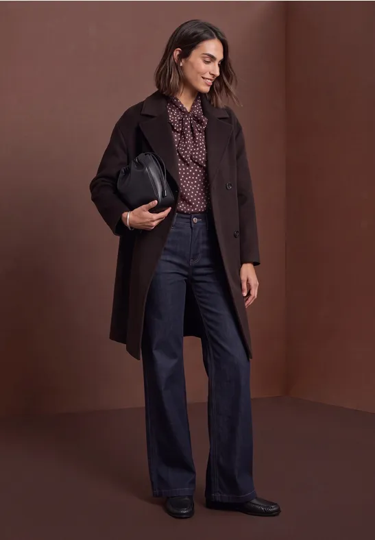 Femme en manteau marron, chemisier à motifs et jean large, tenant un sac noir.