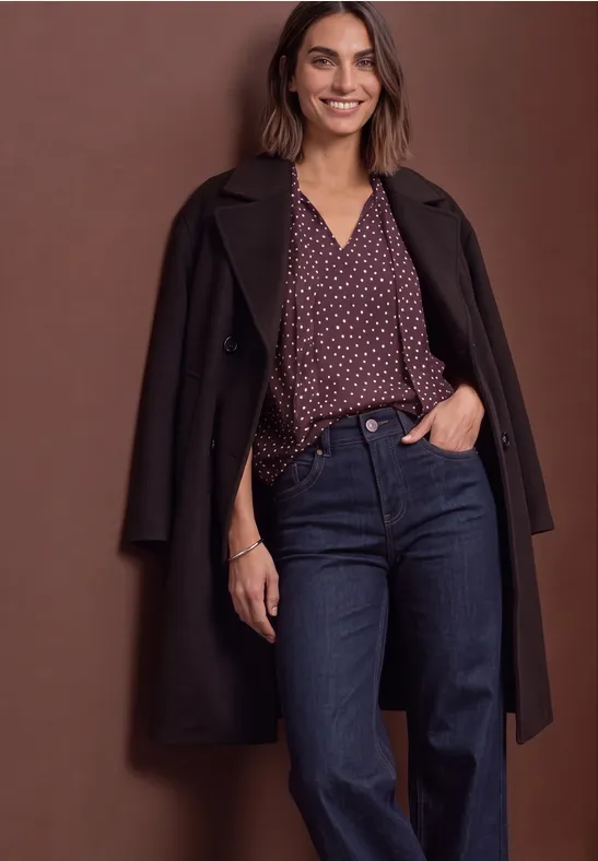 Femme souriante en manteau sombre, chemisier violet à pois et jean bleu, posant devant un fond marron.