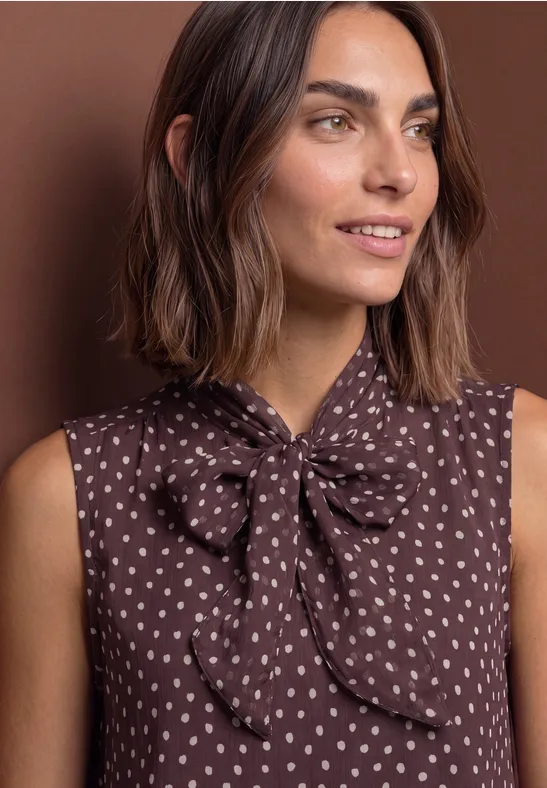 Femme en blouse marron sans manches à motif de coeurs blancs et nœud au col, fond brun.
