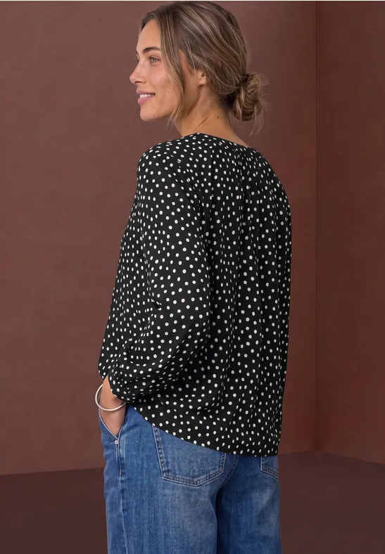 Frau im Profil trägt schwarze gepunktete Bluse und Jeans, lächelnd.