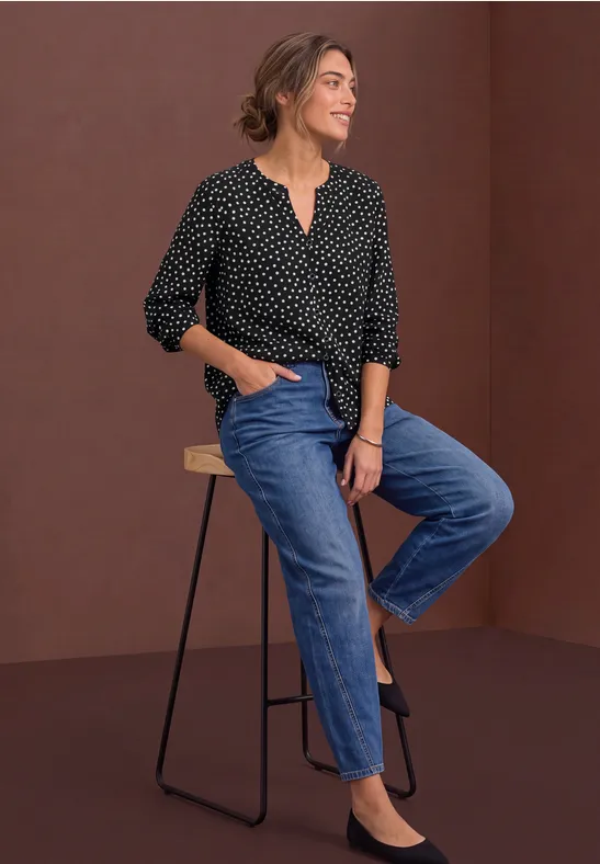 Frau in schwarzer, gepunkteter Bluse und blauen Jeans sitzt lächelnd auf einem Hocker vor braunem Hintergrund.