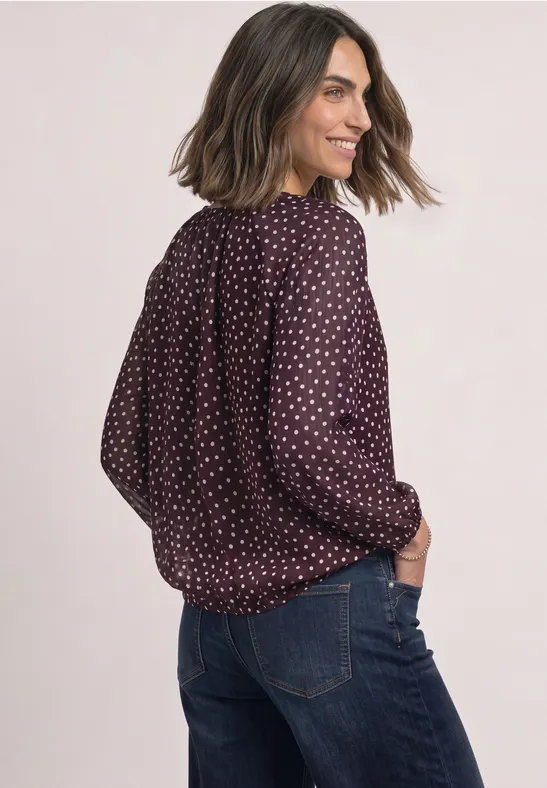 Eine Frau trägt eine bordeauxrote, gepunktete Bluse und Jeans, schaut seitlich über die Schulter.