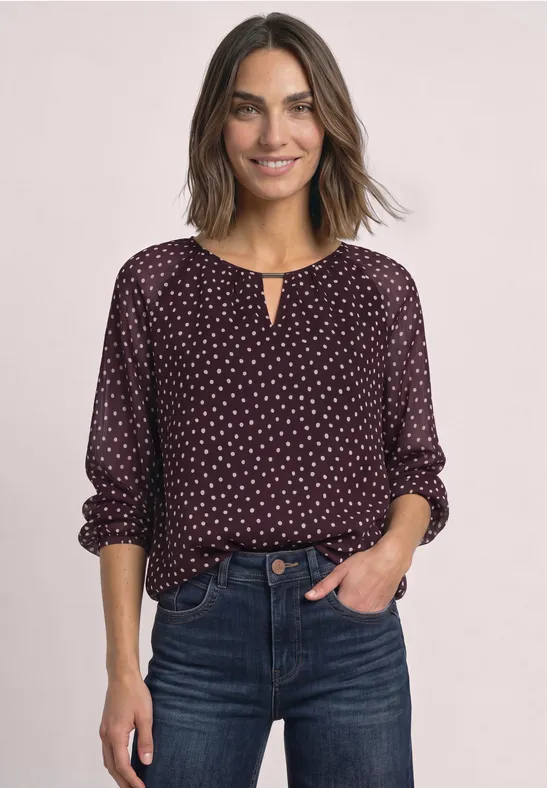 Eine lächelnde Frau mit kastanienbrauner Bluse mit weißen Punkten und dunklen Jeans vor hellem Hintergrund.