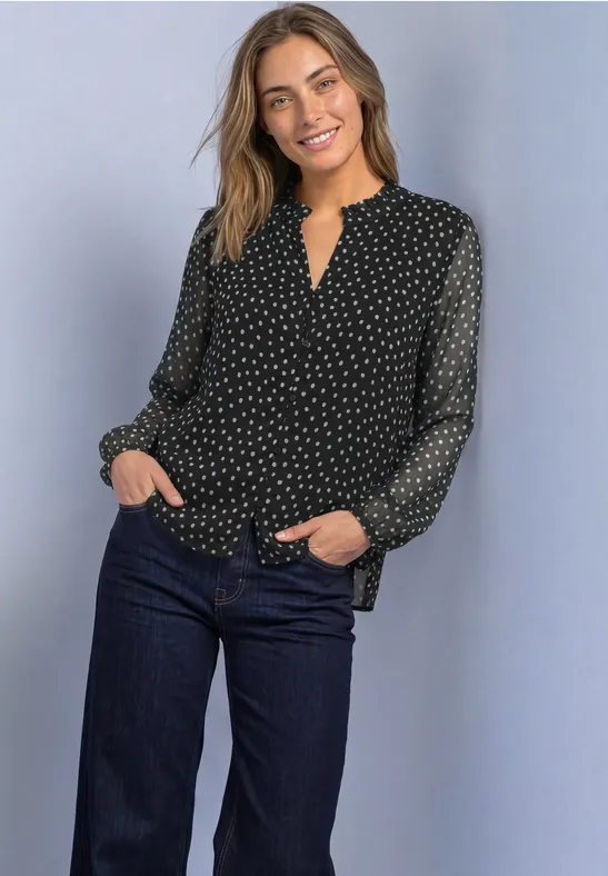 Eine Frau steht lächelnd, trägt eine schwarze, gepunktete Bluse und dunkle Jeans.