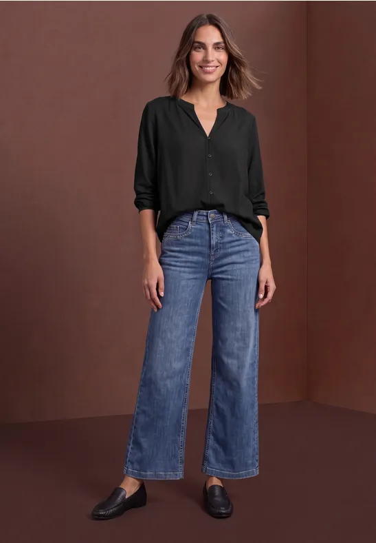 Frau in schwarzer Bluse und blauen Jeans steht vor braunem Hintergrund.