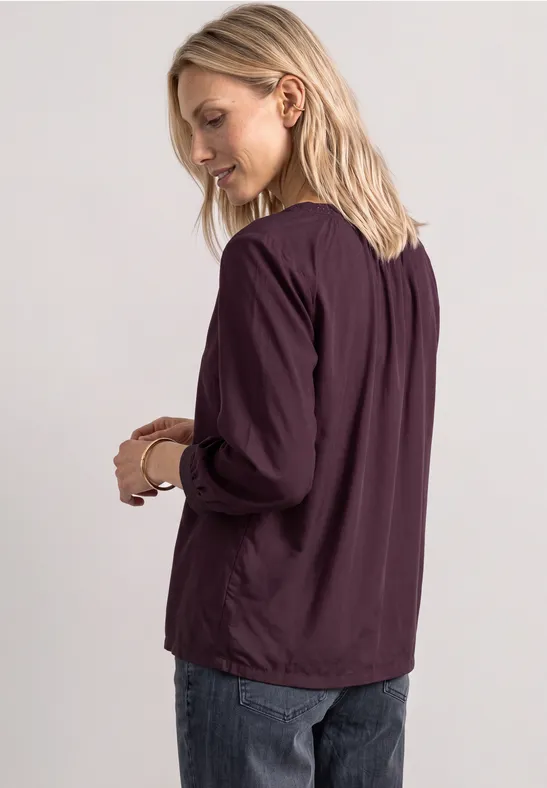Eine blonde Frau trägt eine auberginefarbene Bluse und Jeans, steht seitlich und schaut nach unten.
