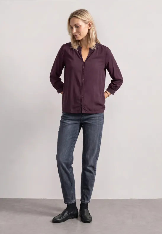 Eine Frau trägt eine lila Hemdjacke, Jeans und schwarze Stiefeletten, steht vor neutralem Hintergrund.