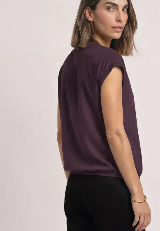 Une femme porte un top sans manches violet et un pantalon noir, regarde par-dessus l'épaule.