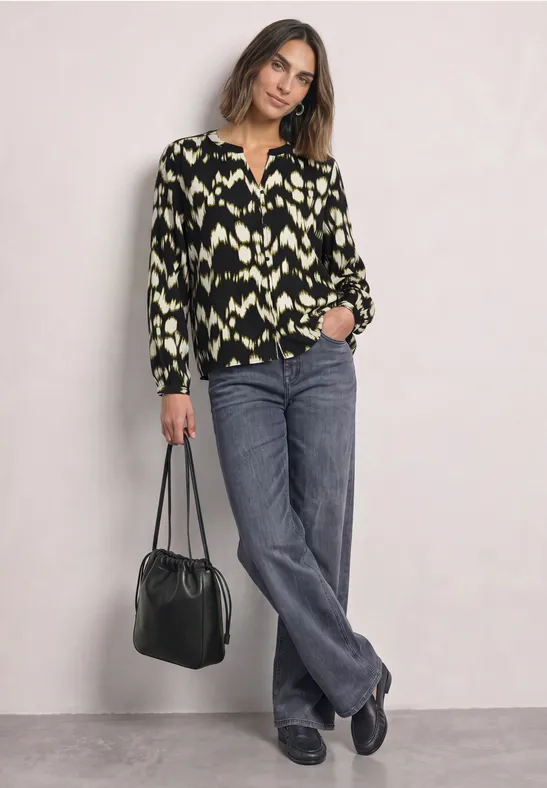 Eine Frau trägt eine gemusterte Bluse, graue Jeans und schwarze Schuhe, hält eine schwarze Tasche.