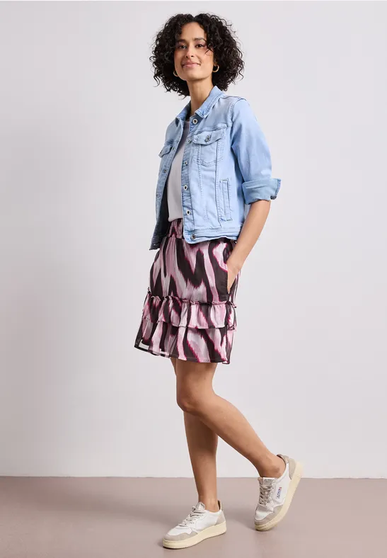 Femme portant une veste en jean bleu clair, une jupe à motif et des baskets devant un fond neutre.