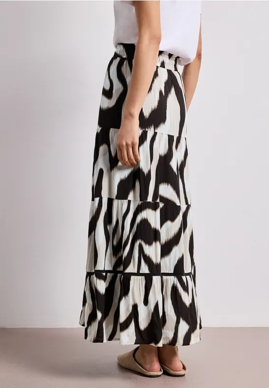 Seitansicht einer Frau in weißem Top und langem Maxirock mit schwarz-weißem Zebra-Muster, getragen mit flachen Sandalen.