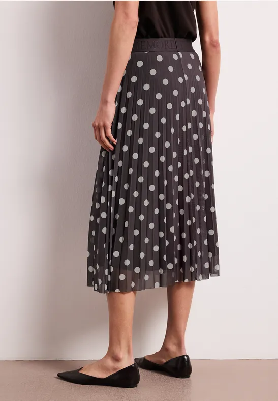 Femme portant une jupe midi plissée noire à pois blancs, associée à des ballerines noires simples.