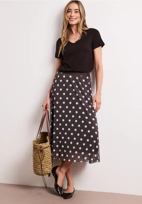 Femme en t-shirt noir et jupe midi à pois, portant un sac en paille et des ballerines sur fond clair.