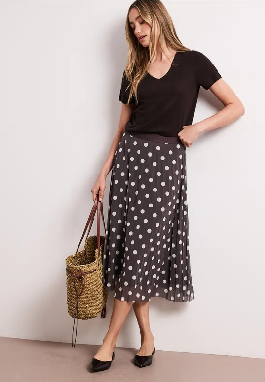 Femme en t-shirt noir, jupe midi à pois blancs, sac en paille et mules noires.