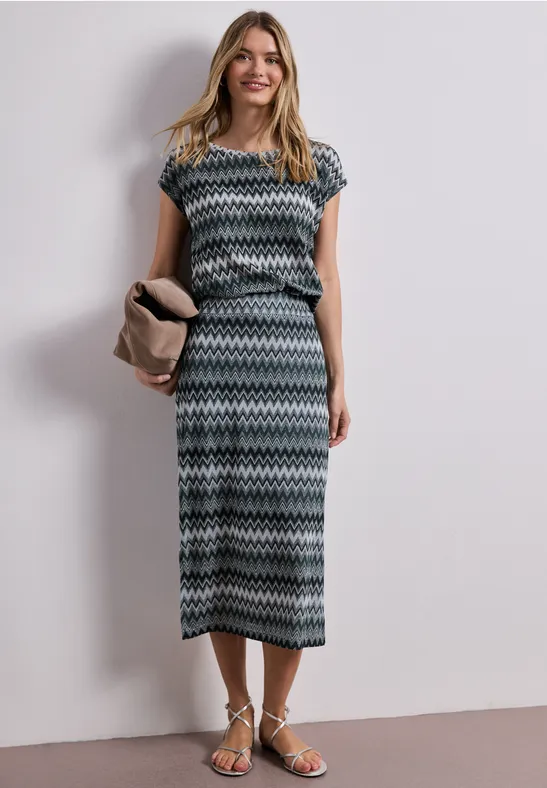 Vrouw in een midi-jurk met korte mouwen en zigzagprint, platte sandalen en clutch in neutrale kleur.