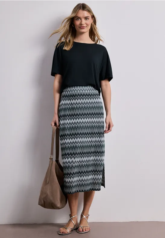 Een vrouw draagt een zwart T-shirt, een midi-rok met patroon, sandalen en houdt een grote tas.