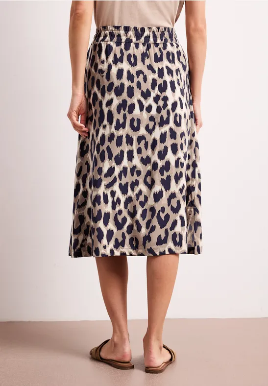 Achteraanzicht van een persoon in een midirok met luipaardprint en sandalen tegen een neutrale achtergrond.