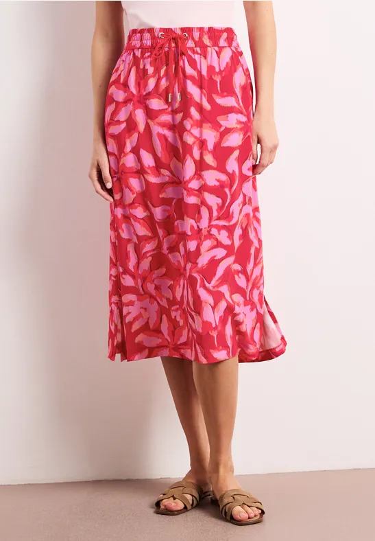 Rode midi-rok met roze bladprint, soepel vallend, gecombineerd met platte sandalen op een lichte achtergrond.