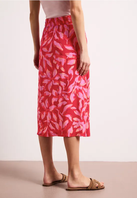 Achteraanzicht van een vrouw in een rode midirok met bladmotief en beige sandalen.