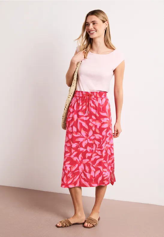 Een vrouw draagt een roze t-shirt, een rode rok met bladprint, sandalen en een strooien tas.