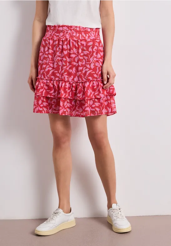 Unterkörper einer Person in weißem Shirt und Sneakers, rotem Rock mit weißem Blattmuster und Rüschen.