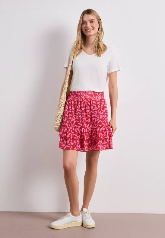 Frau in weißem T-Shirt, rotem gemustertem Rock und weißen Sneakern mit Strohtasche vor hellem Hintergrund.