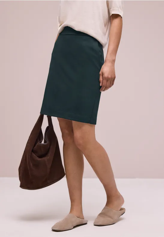 Vrouw in witte blouse en groene rok met bruine tas en beige instappers voor een neutrale achtergrond.