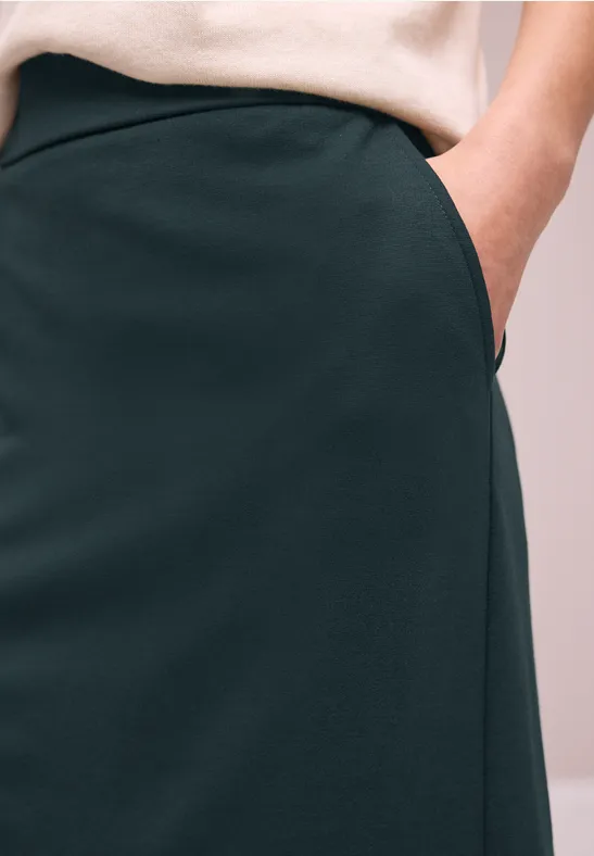 Close-up van een donkergroene rok met zijzak en hand erin, gecombineerd met een wit bovenstuk.