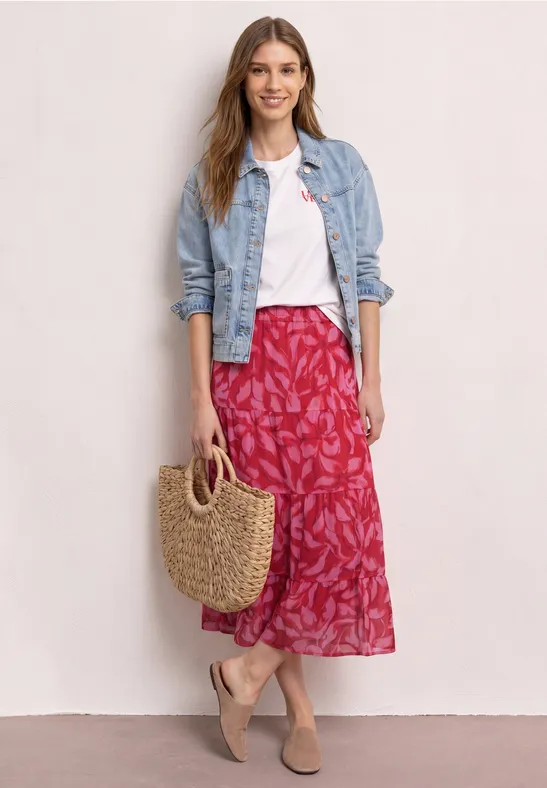 Vrouw in wit shirt, spijkerjasje, rood bloemenrok en rietentas.