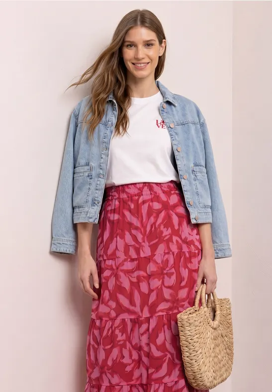 Vrouw in witte T-shirt, spijkerjas en een roze, gebloemde rok met strooien mandje voor neutrale achtergrond.