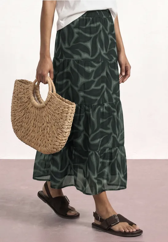 Groene midi-rok met bladdessin, bruine sandalen en een ronde strozak in de hand.