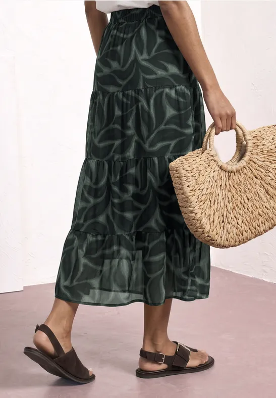Groene maxirok met bladprint, zwarte sandalen en een ronde rieten tas in de hand.